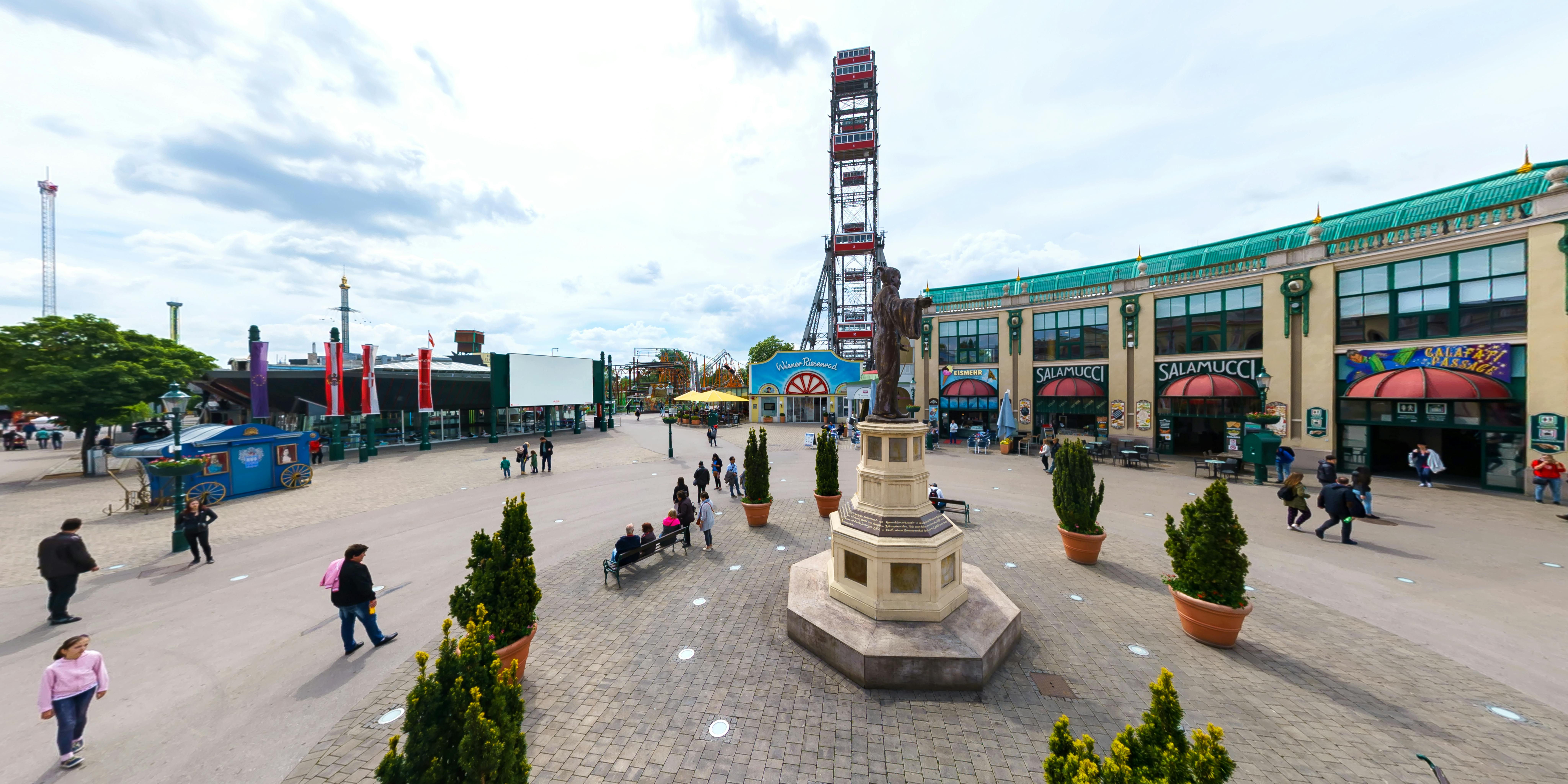 Socha stojí na náměstí obklopeném květináči s lavičkami obsazenými lidmi. V pozadí je ruské kolo a obchody.