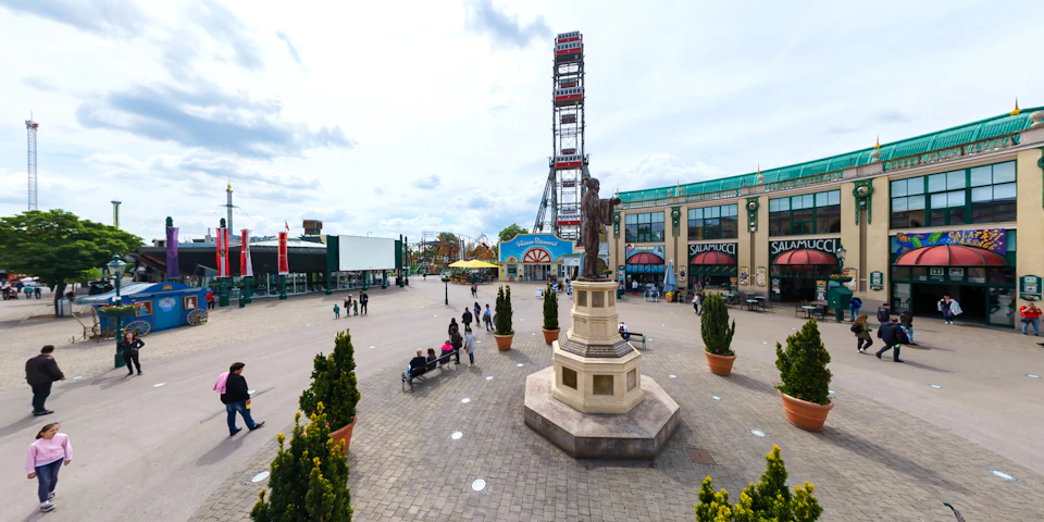 Public square with a statue in the center, potted plants, benches, and buildings. A tall amusement park ride is in the background.