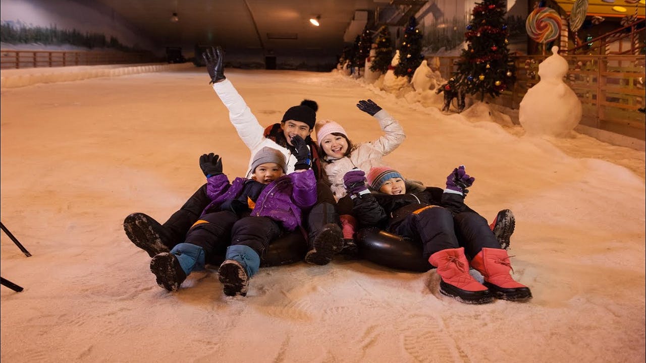 Família de quatre, vestits amb roba d'hivern, asseguts sobre tubs de pneumàtics en un vessant cobert de neu, saludant i somrient a la càmera.