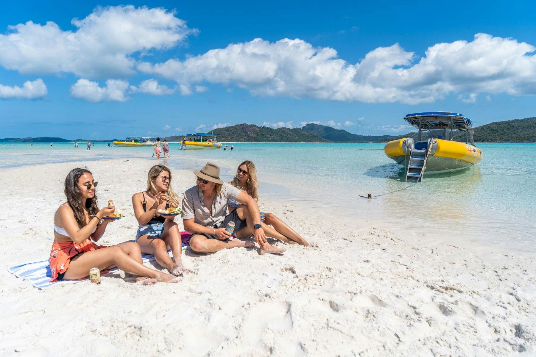 Four people sitting on a sandy beach, eating and relaxing under a sunny sky with boats and an island in the background.