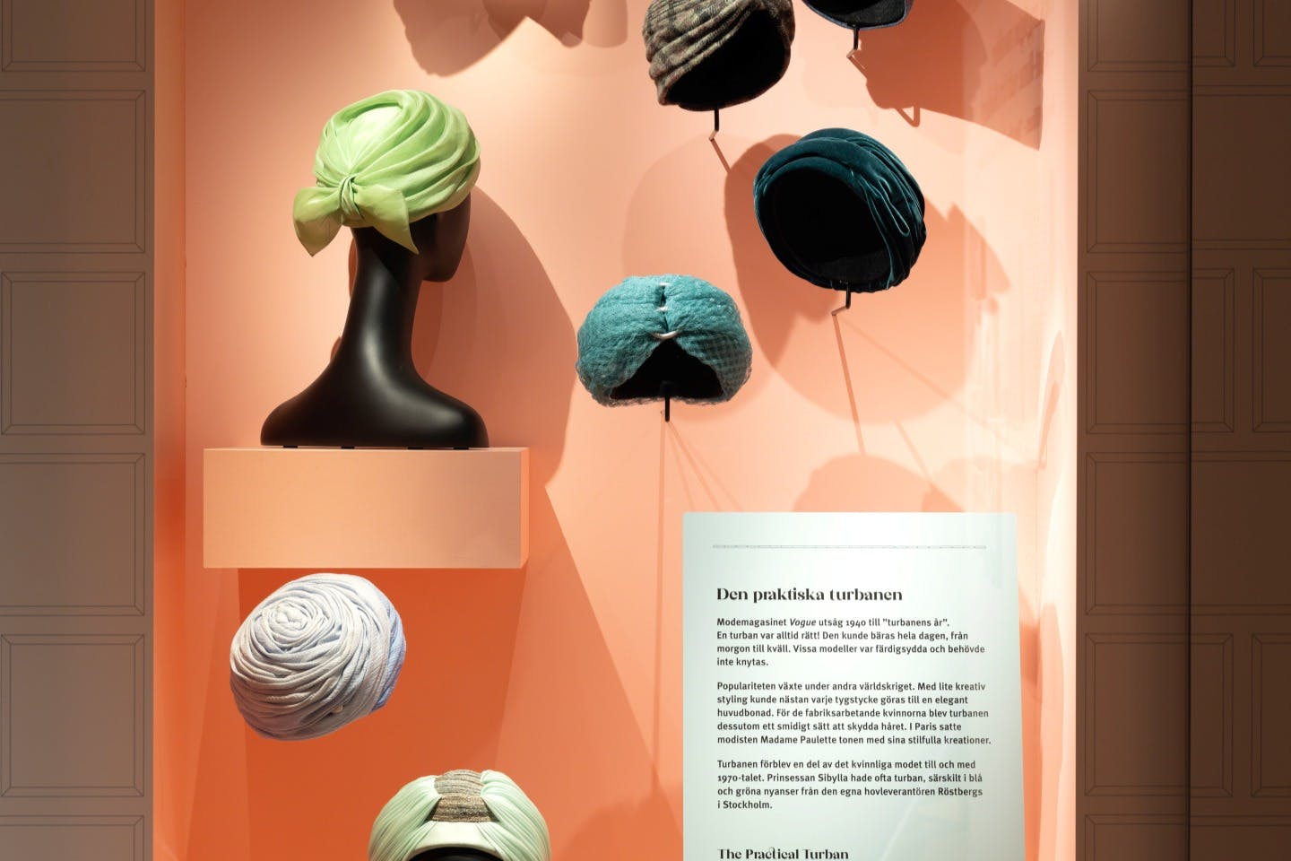 Esposizione di vari turbanti colorati su manichini e supporti su una parete color pesca, con una targa descrittiva in svedese e in inglese.