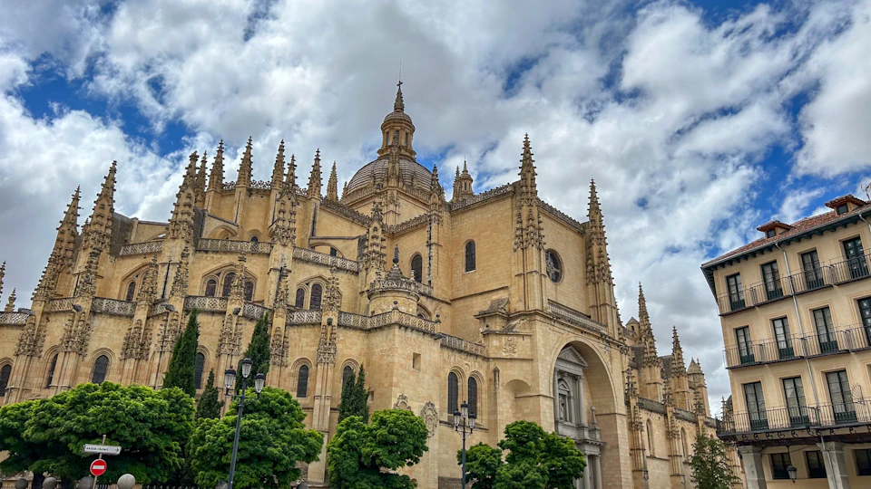 Gothic cathedral with spires and a large dome, surrounded by green trees against a partly cloudy sky.