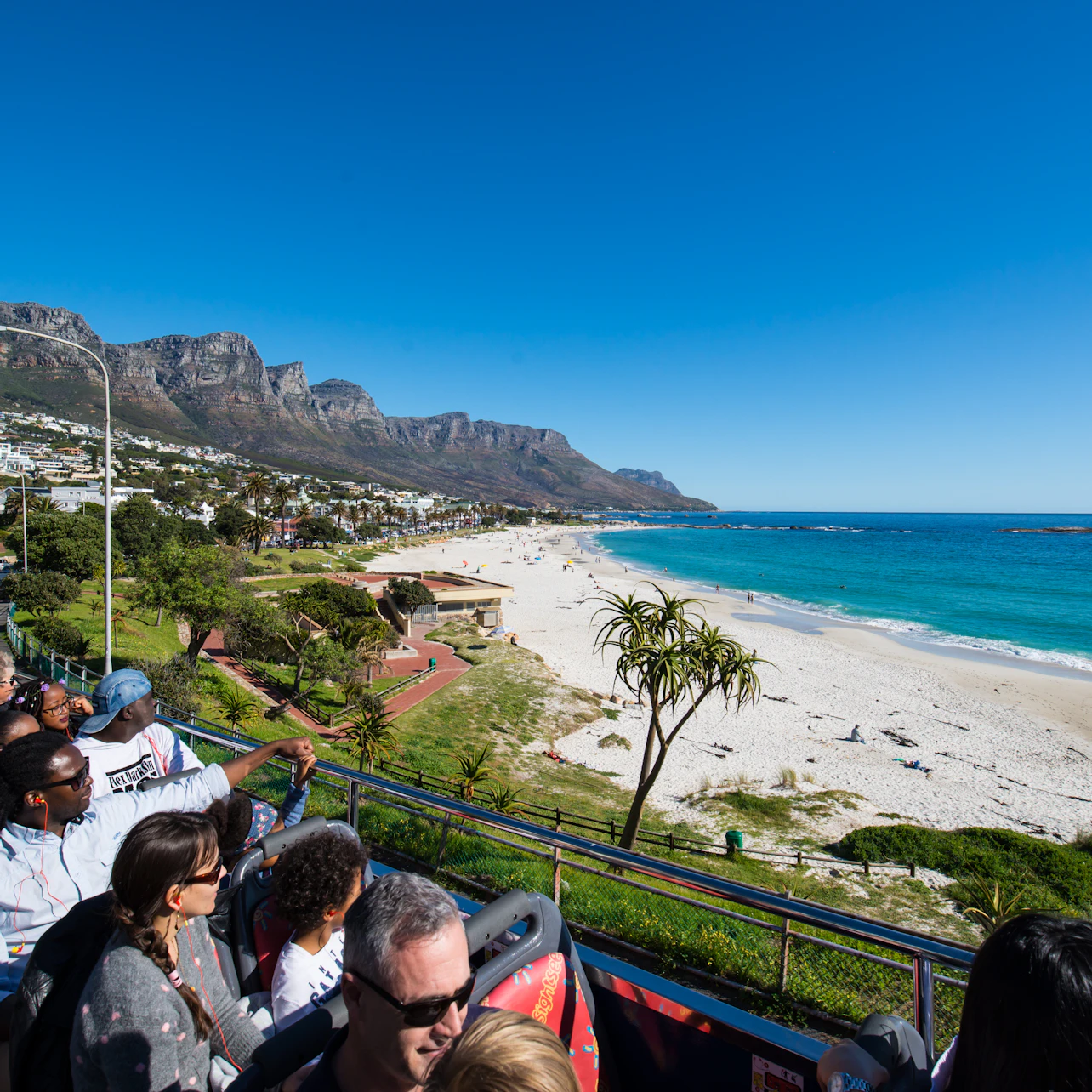 Tourists in an open-top bus enjoy a scenic view of a coastal beach, mountains, and clear blue sky.