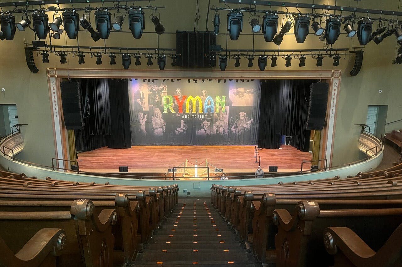 Teatro vuoto con sedili in legno rivolti verso un palco. Sullo sfondo compare la scritta "Ryman Auditorium" con immagini di artisti.
