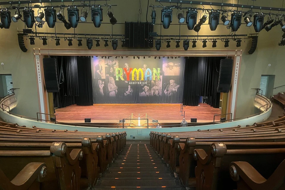 Empty theater with wooden seats facing a stage. A backdrop displays "Ryman Auditorium" with images of performers.