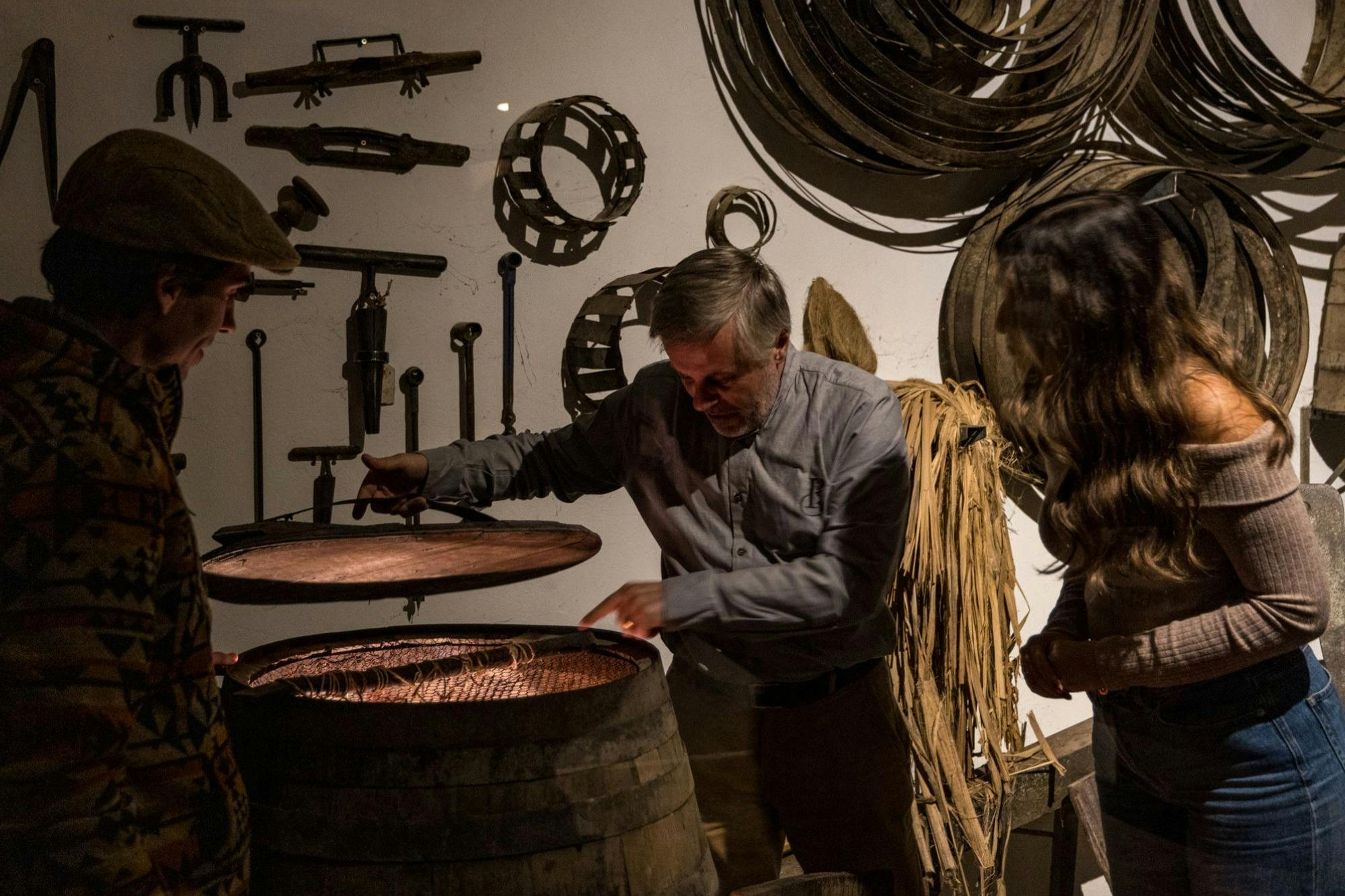 A man demonstrating the contents of a barrel to two people in a rustic setting with tools and materials on the wall.