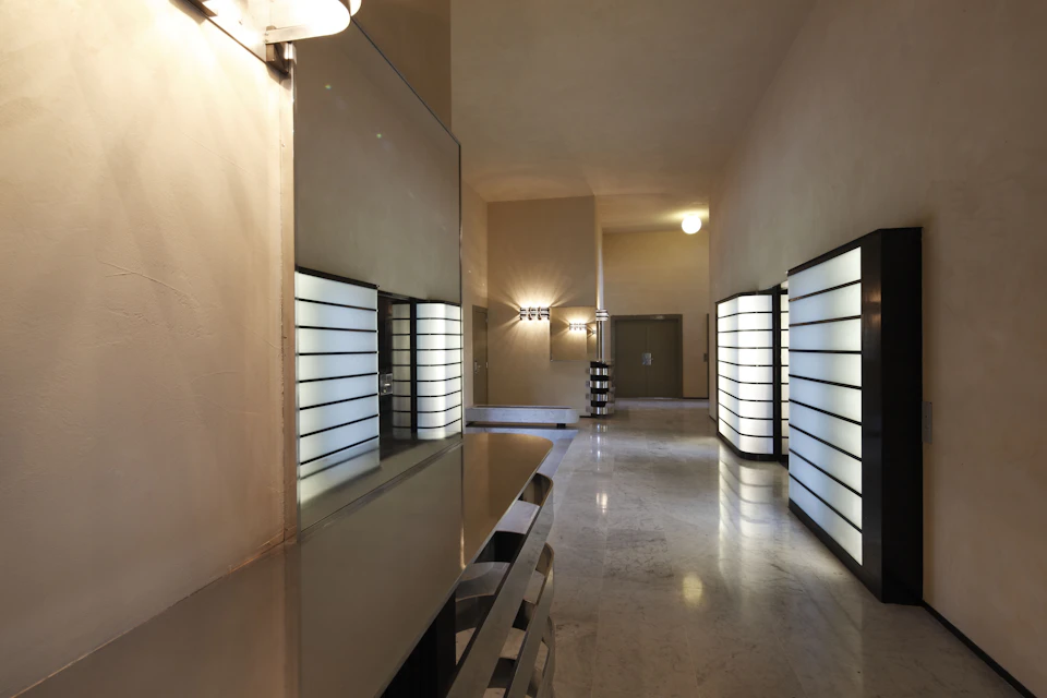 A well-lit, minimalist hallway with reflective surfaces, modern light fixtures, and large, illuminated wall panels on both sides.
