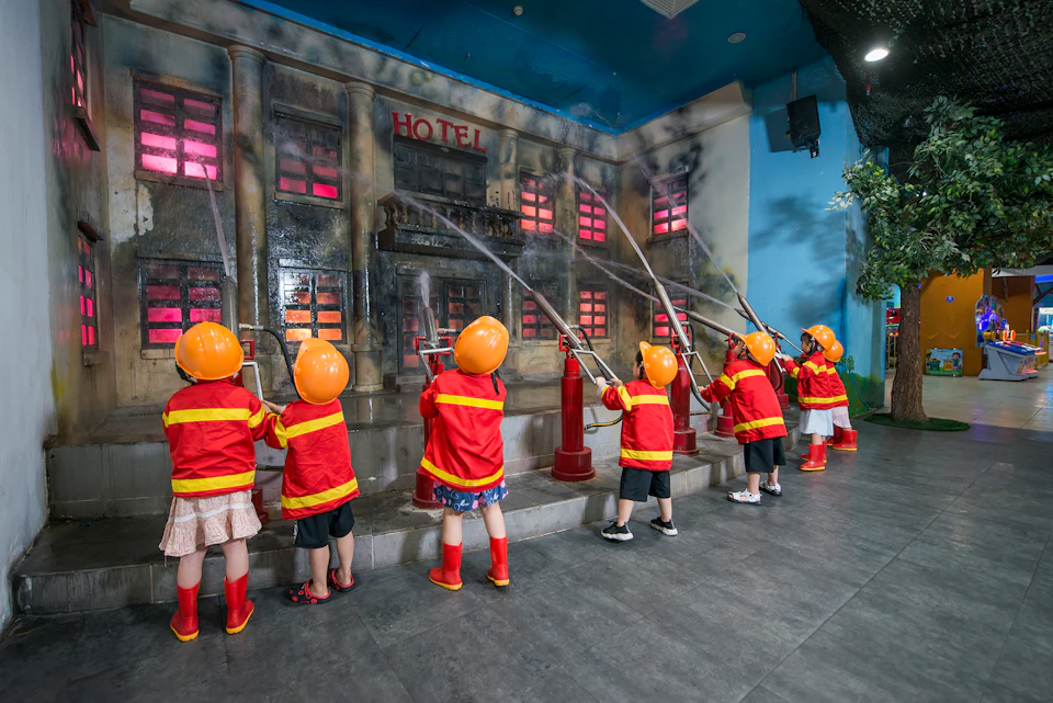Children wearing firefighter costumes spray water at a simulated burning building with red-lit windows.
