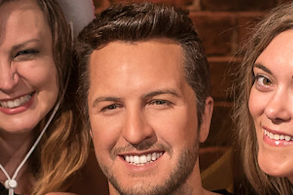 A man with a guitar is surrounded by four smiling women wearing costumes, including cowboy hats and tiaras, against a brick wall.