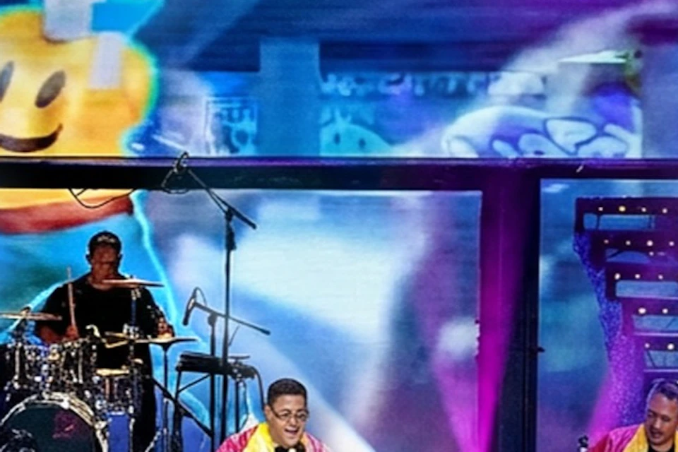 A lively musical performance onstage with colorful lights, a band, and visuals, featuring animated characters and a Rubik's Cube.