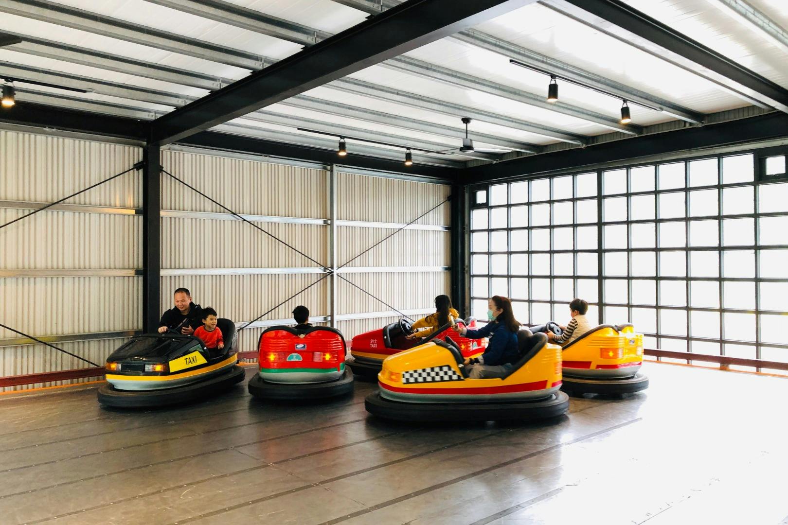 People riding colorful bumper cars indoors in a metal-framed building.