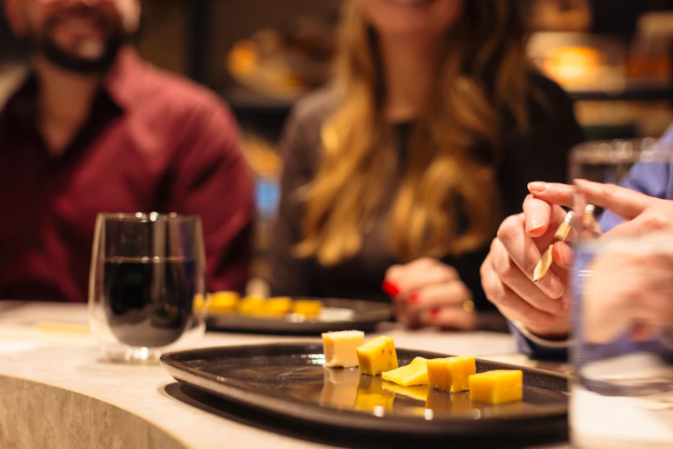 Close-up of cheese cubes on a black plate, with blurred people in the background and a glass of water on the table.