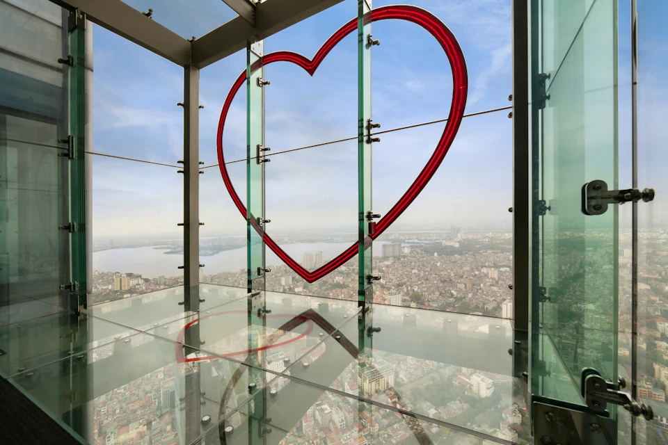 A red heart-shaped outline is attached to glass panels of an observation deck, overlooking a cityscape under a blue sky.