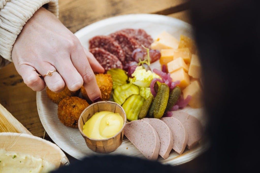 Hand reaching for food on a platter with sliced meats, cheese cubes, pickles, mustard, and fried items.
