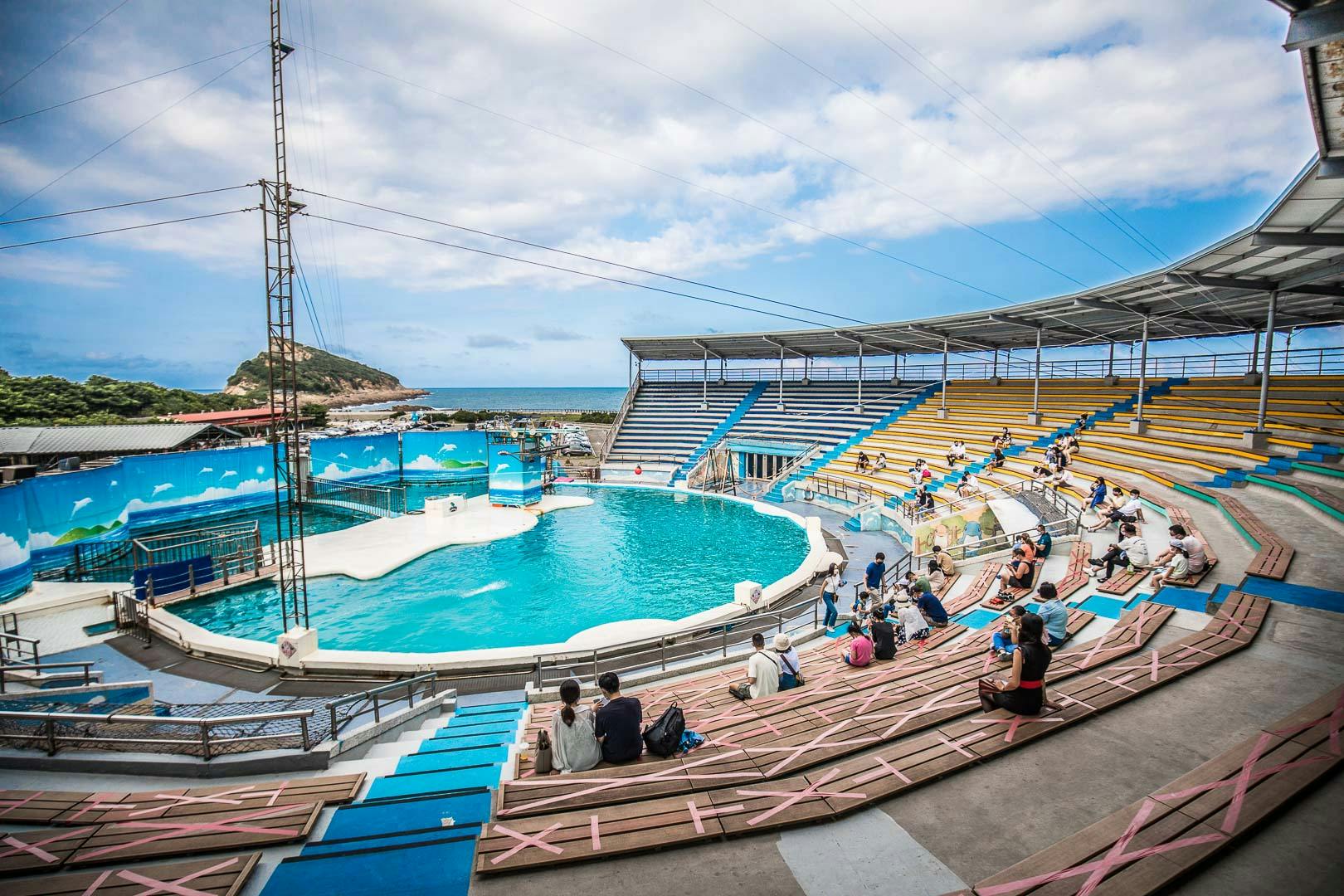 Un amphithéâtre en plein air avec une grande piscine au centre, entouré de gradins, donnant sur un paysage côtier par une journée ensoleillée.