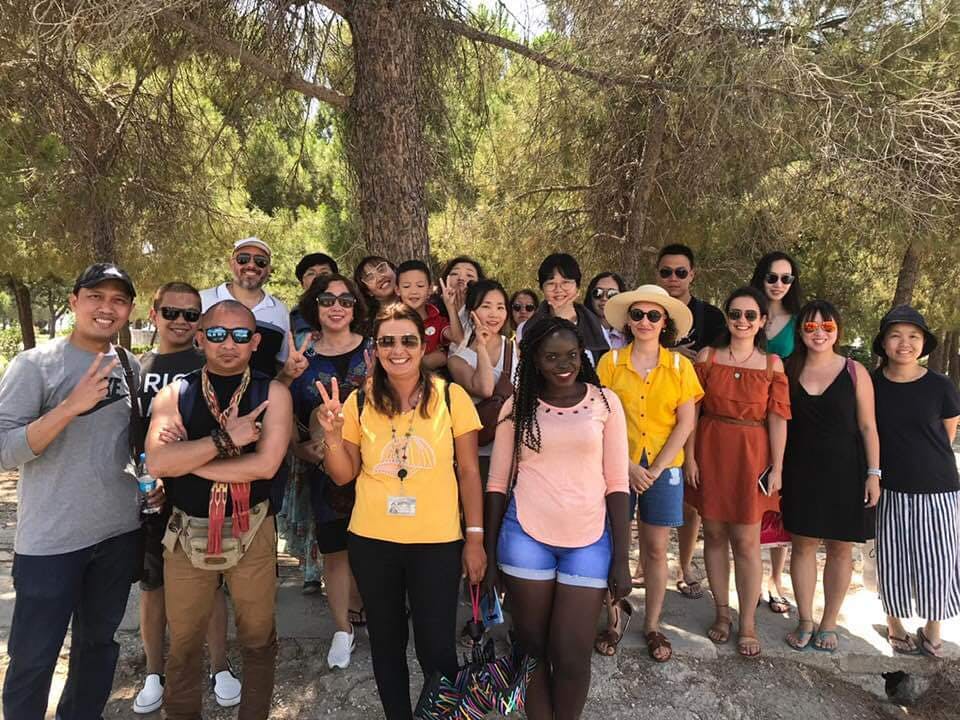 日当たりの良い屋外環境で、木の下でポーズをとる多様な人々のグループ。多くの人が笑顔でピースサインを作っています。