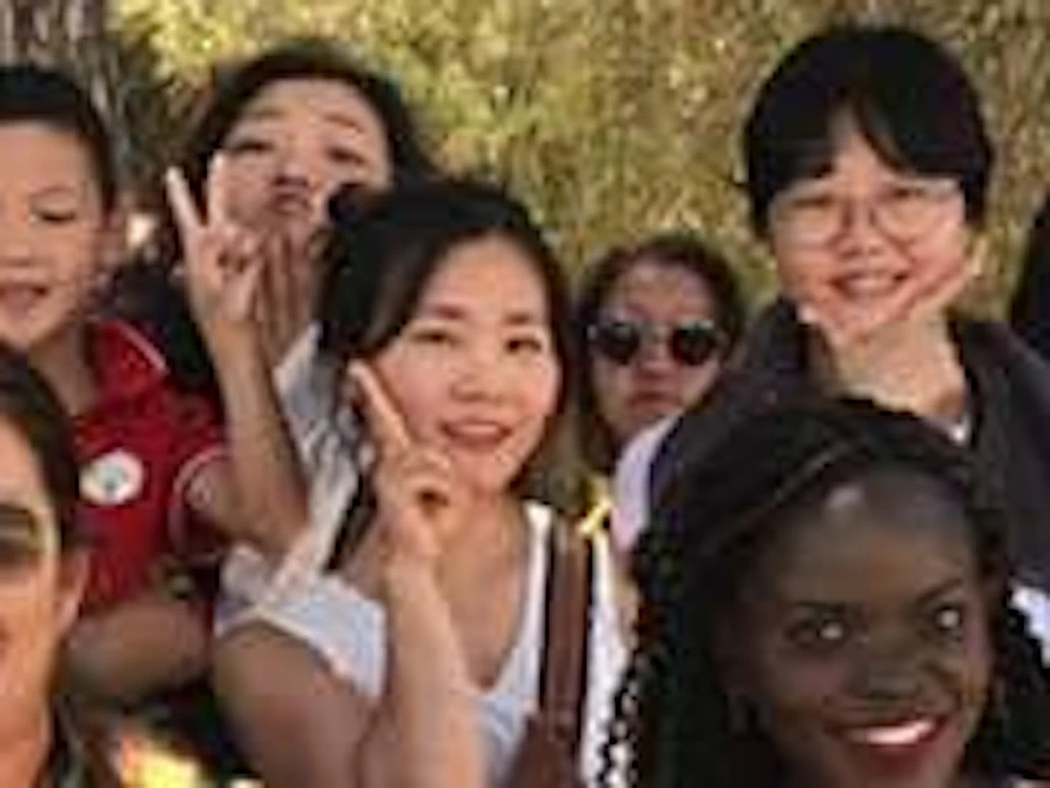 A diverse group of people posing under trees, many smiling and making peace signs, in a sunny outdoor setting.