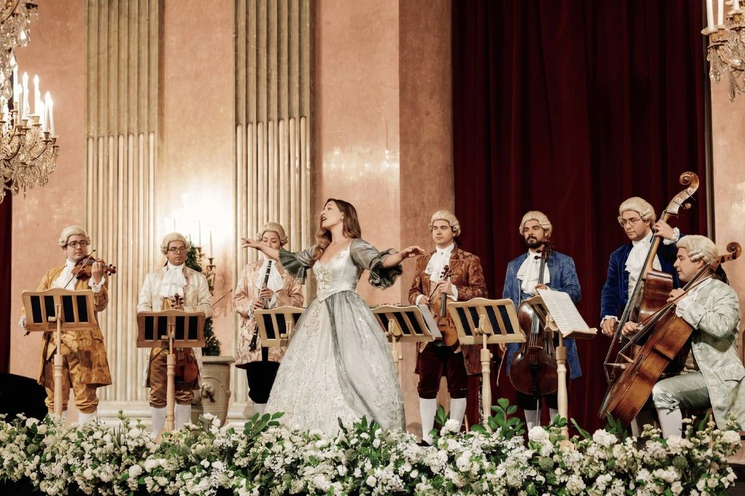A baroque-style ensemble with period costumes performs on stage. A soprano sings in front of musicians playing string instruments.