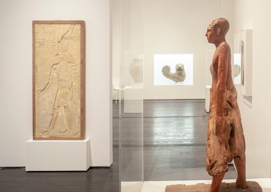 창문을 통해 추상적인 조각을 배경으로 목조 휴머노이드 조각상을 전시하고 있는 박물관 갤러리입니다.