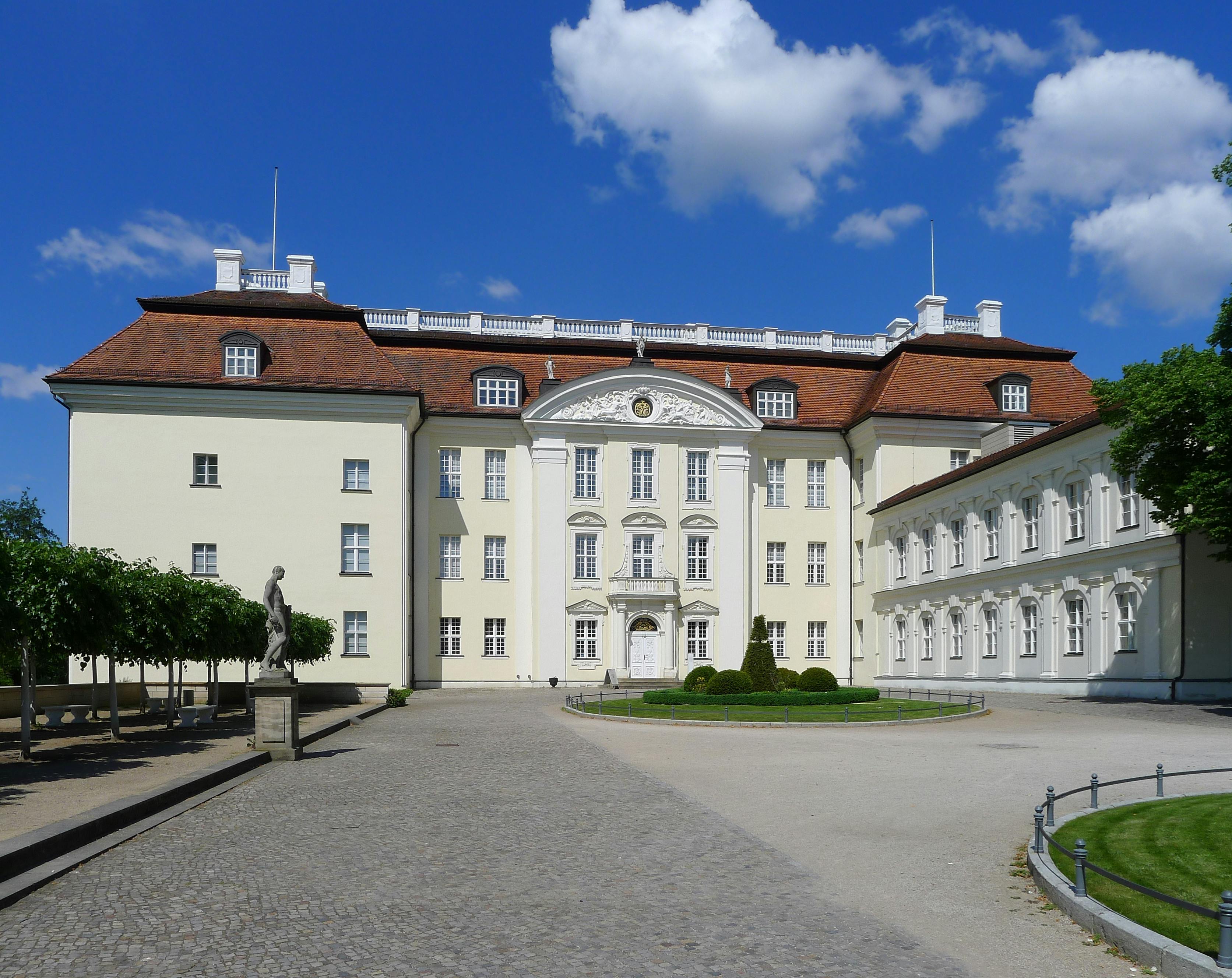 Großes, elegantes Gebäude mit rotem Dach und weißer Fassade, davor ein steinerner Weg und eine Statue, vor einem wolkenlosen blauen Himmel.