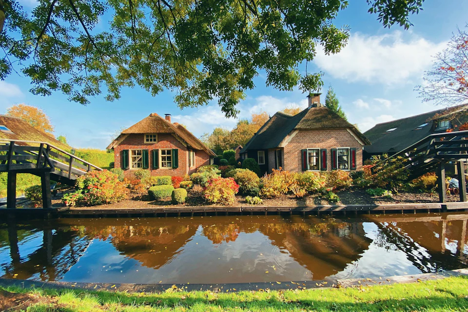 Two thatched-roof cottages surrounded by colorful gardens reflected in a calm canal under a sunny sky.