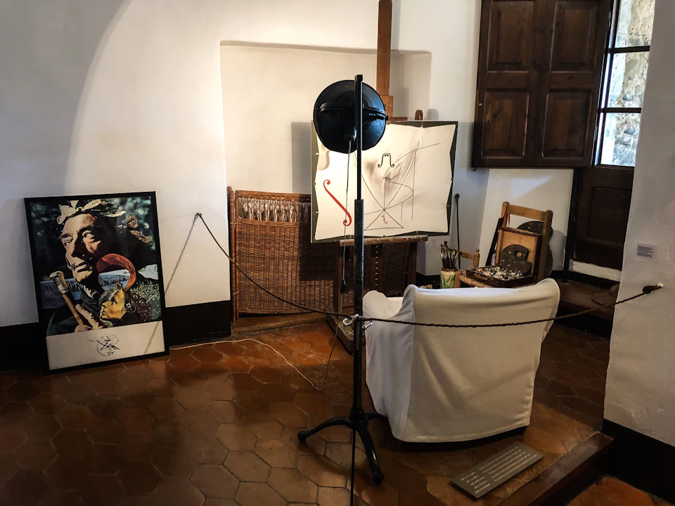 Art studio with a white chair, easel with abstract painting, overhead light, framed picture on the floor, and wooden furniture.