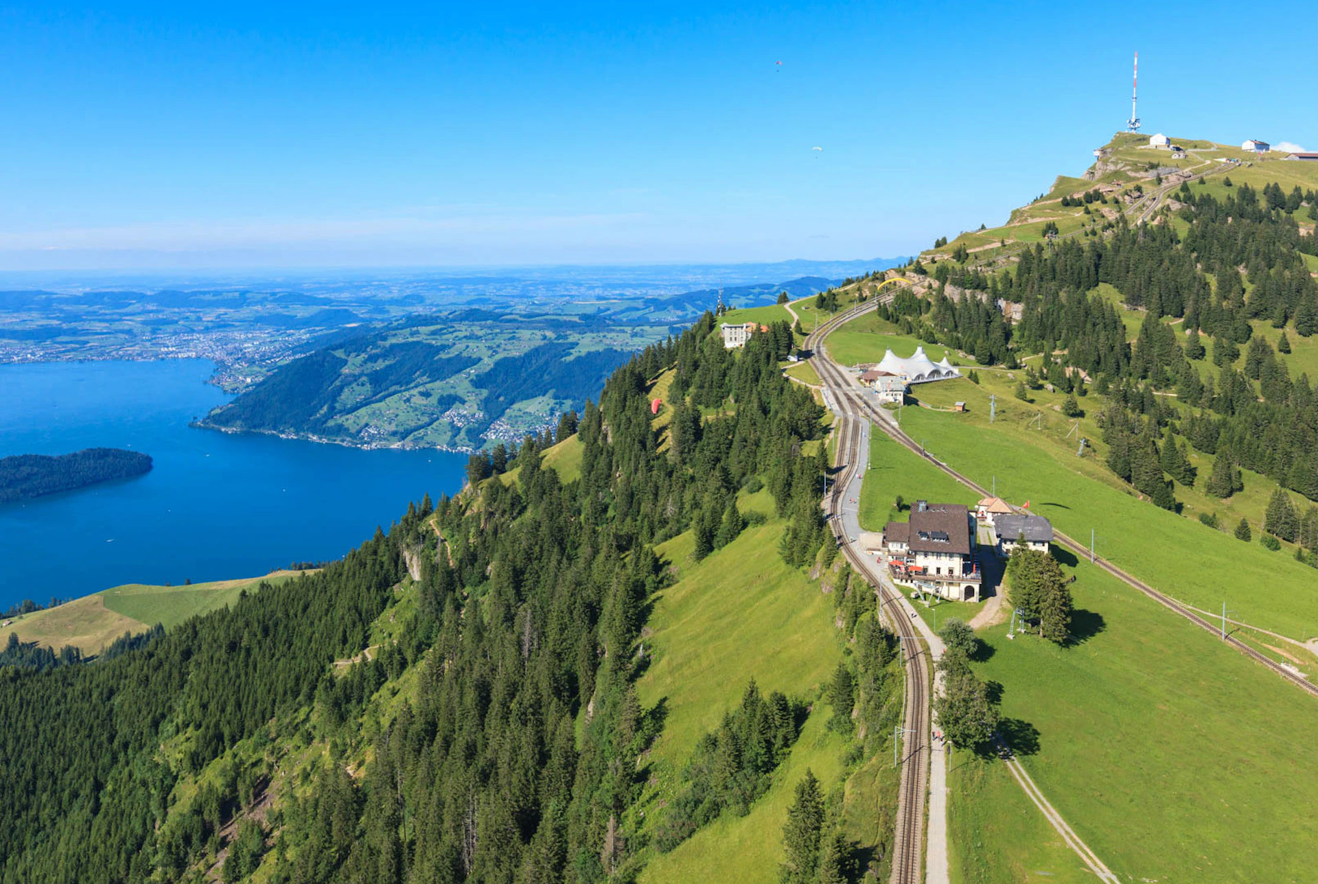A train track winds through a green hilly landscape with trees, buildings, and a blue lake in the background under a clear sky.