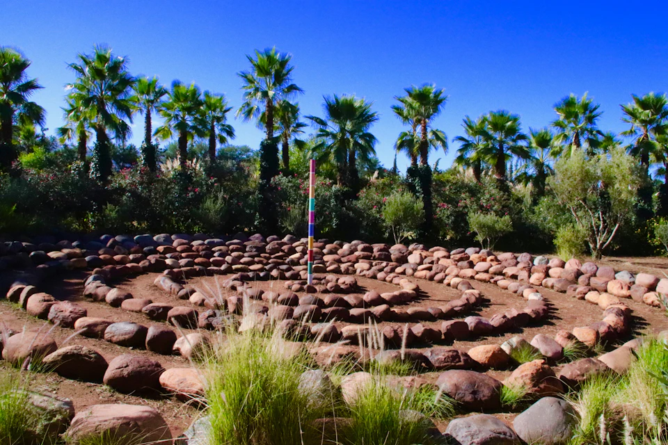 A stone labyrinth with a colorful pole in the center, surrounded by greenery and palm trees under a clear blue sky.