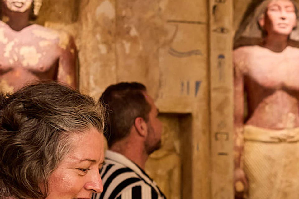 A woman in a museum, smiling while looking at Egyptian artifacts, with another person in the background facing ancient statues.