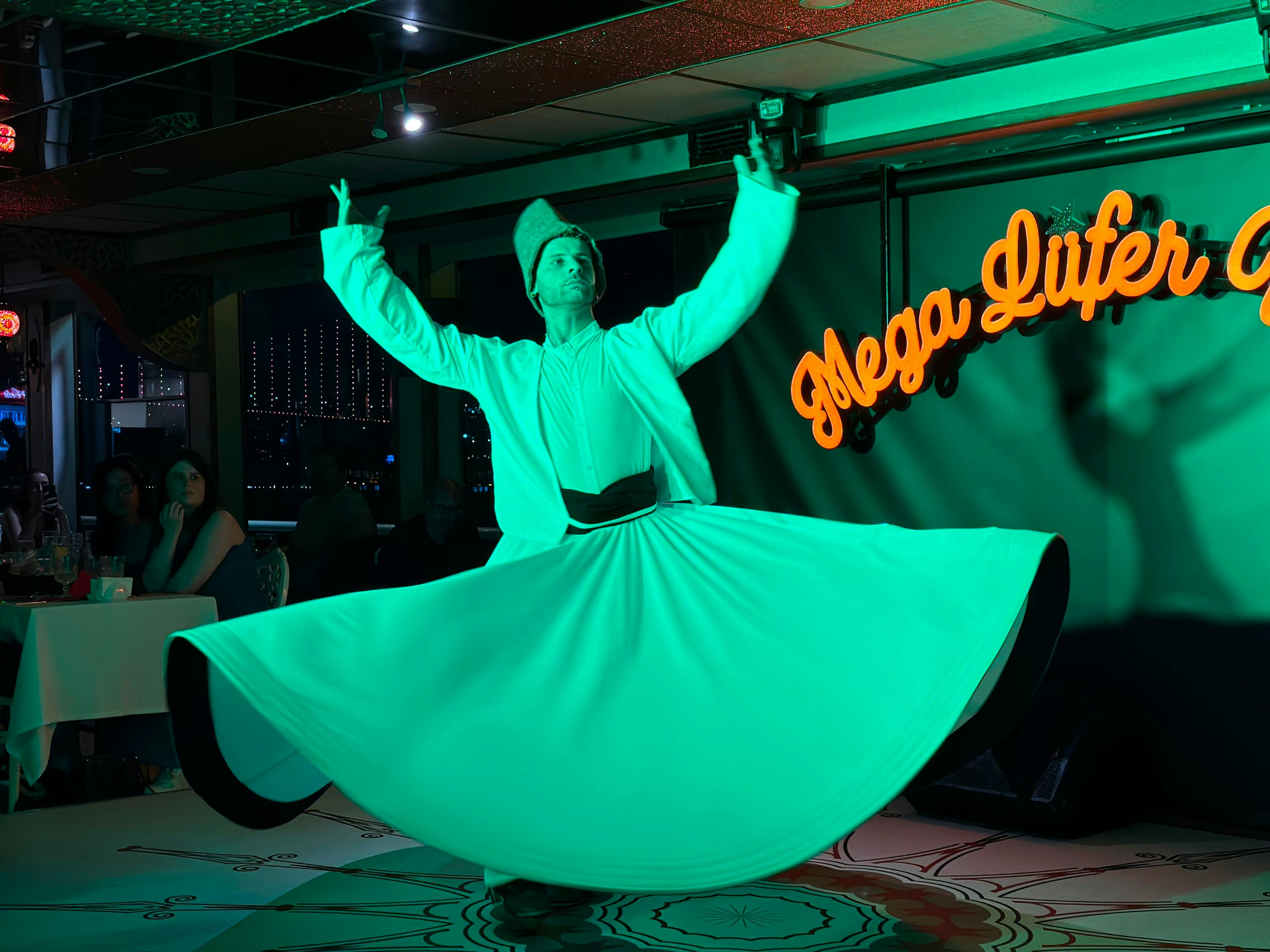 A performer wearing traditional attire spins in a dance on a stage lit in green lighting, with a neon sign in the background.