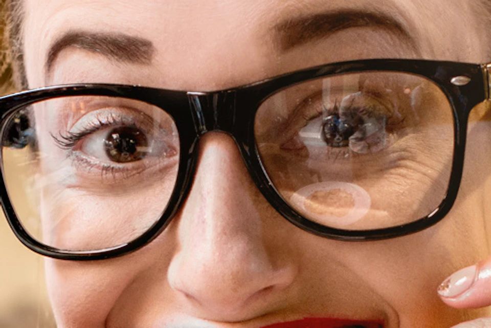 A woman with glasses happily holds a slice of meat in front of her mouth while sitting at a table with various meats and cheeses.