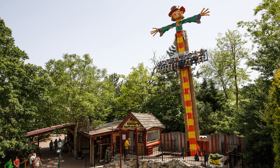 People riding a vertical drop amusement ride with a scarecrow at the top, surrounded by trees and a small building at the base.