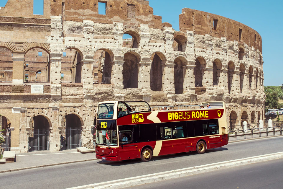 A red and yellow double-decker tour bus labeled 'Big Bus Rome' parked in front of the Colosseum in Rome on a sunny day.