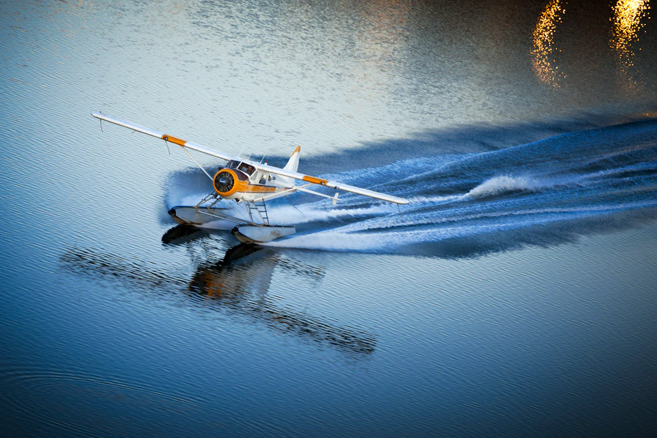 A yellow and white seaplane skims across calm water, creating a wide trail of ripples behind it.