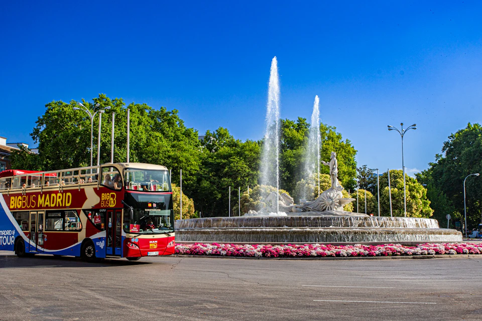 A double-decker tour bus near a fountain with a statue and water jets, surrounded by flowers and greenery under a clear blue sky.