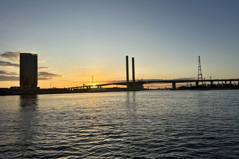 A bridge with two tall pillars spans a river at sunset, with a clear sky and some distant structures visible.