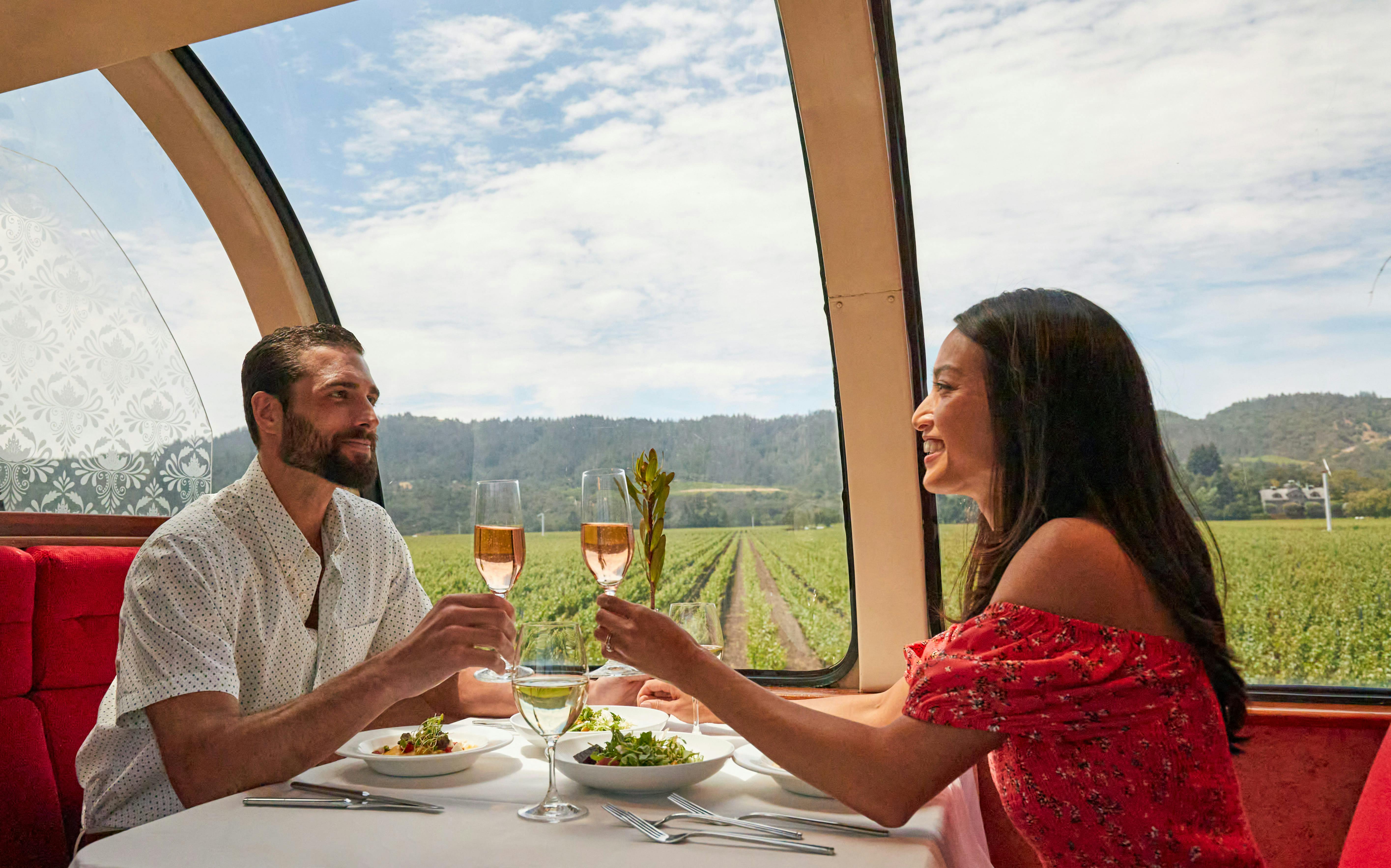 A woman in a red dress holding a glass of wine, sitting at a table with a salad, enjoying a scenic view of vineyards.