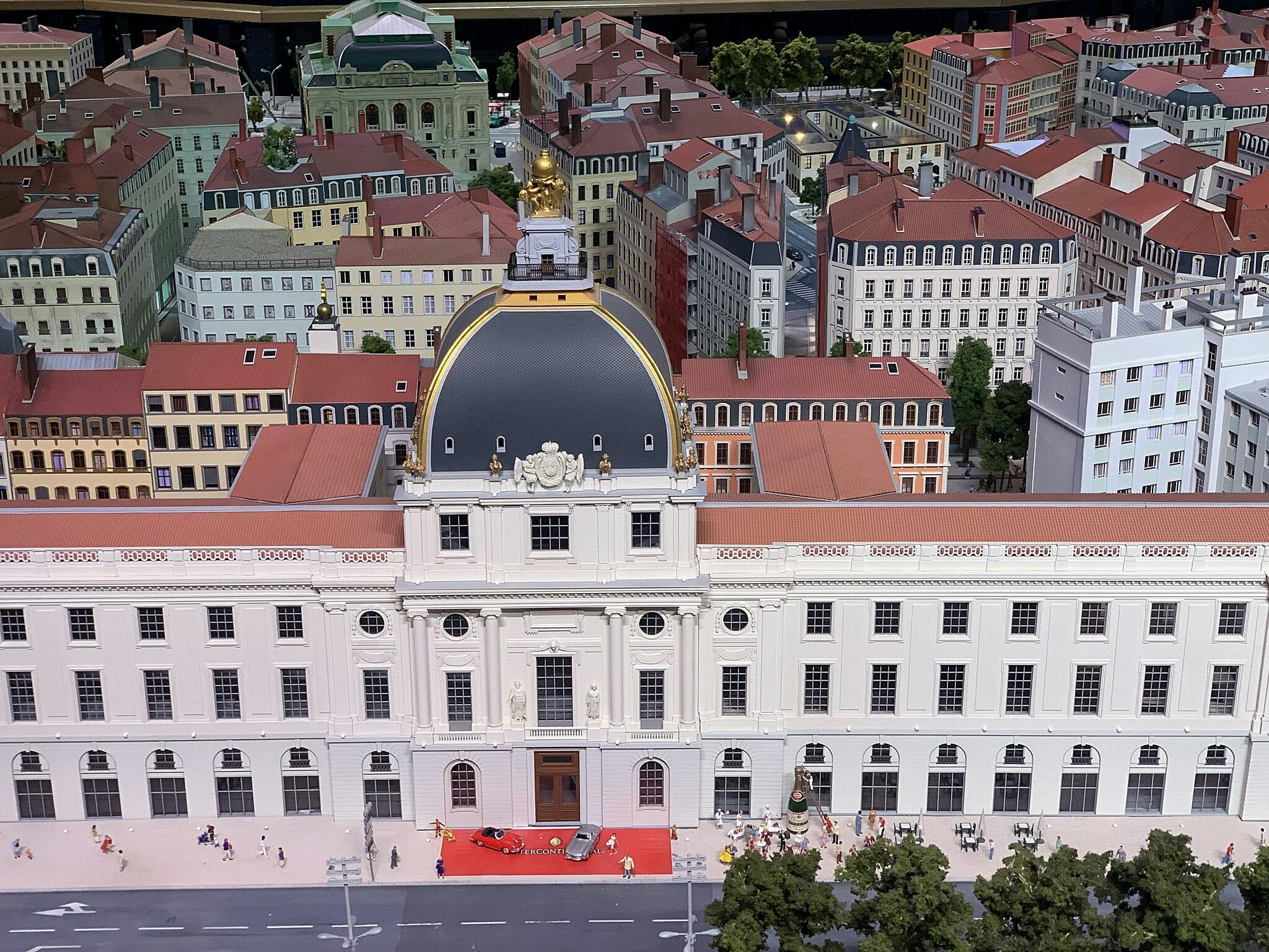 Model of a European town with a prominent classical building, red-roofed structures, and small figurines on streets and sidewalks.