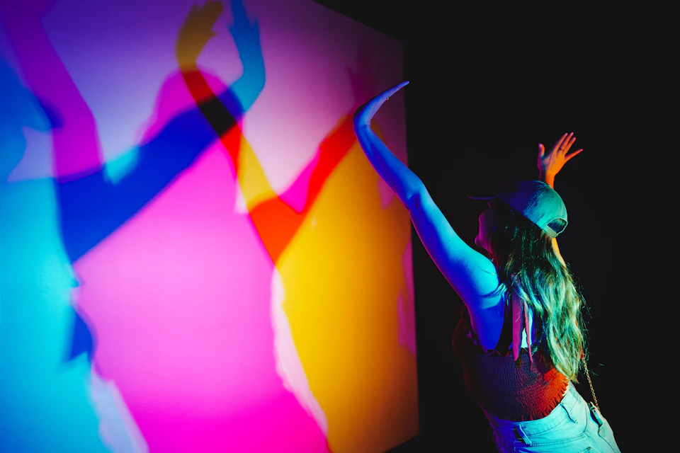 A person wearing a hat interacts with colorful shadows on a wall in a dimly lit space.