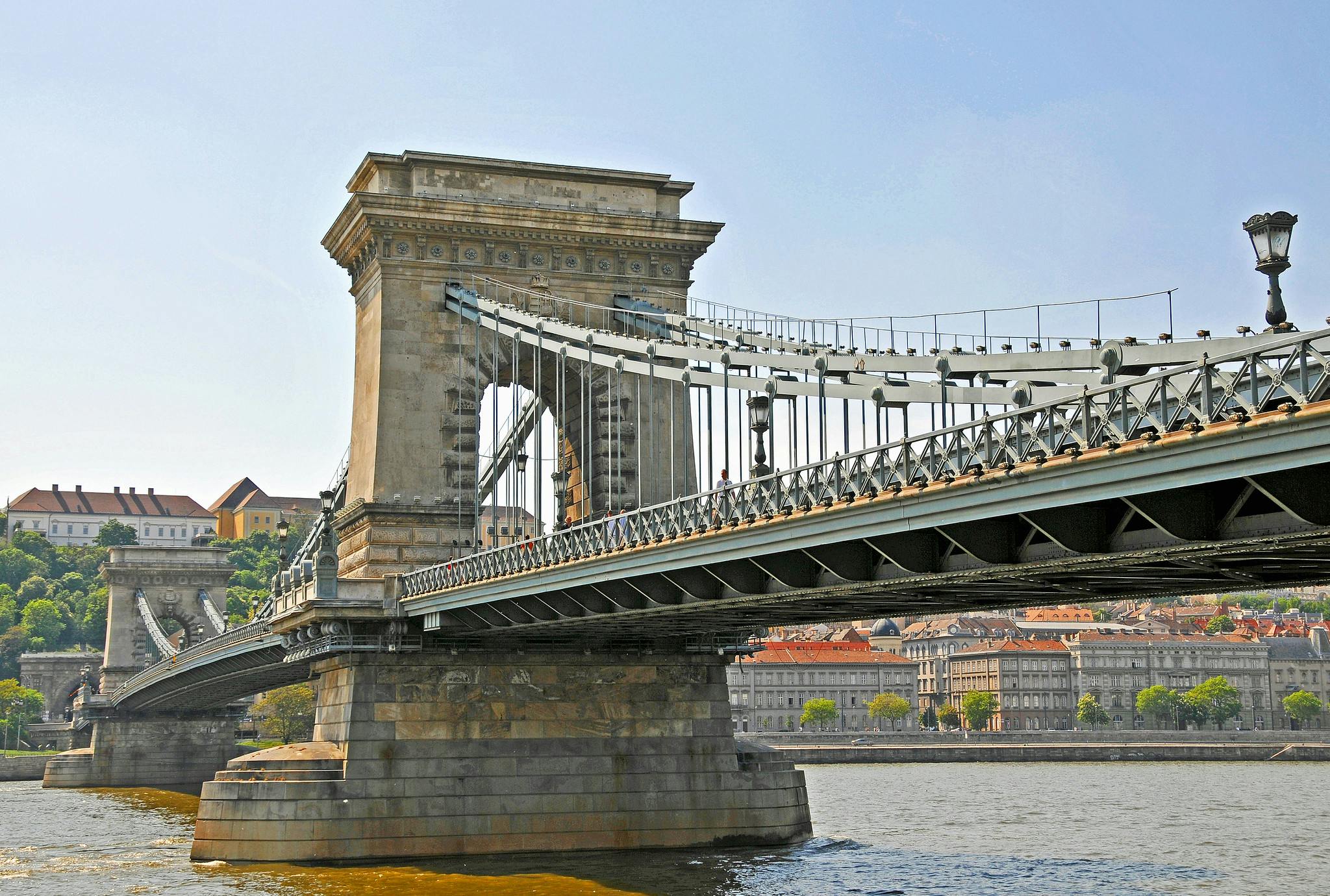 A stone and steel suspension bridge spans across a river with buildings and greenery visible in the background under a clear sky.