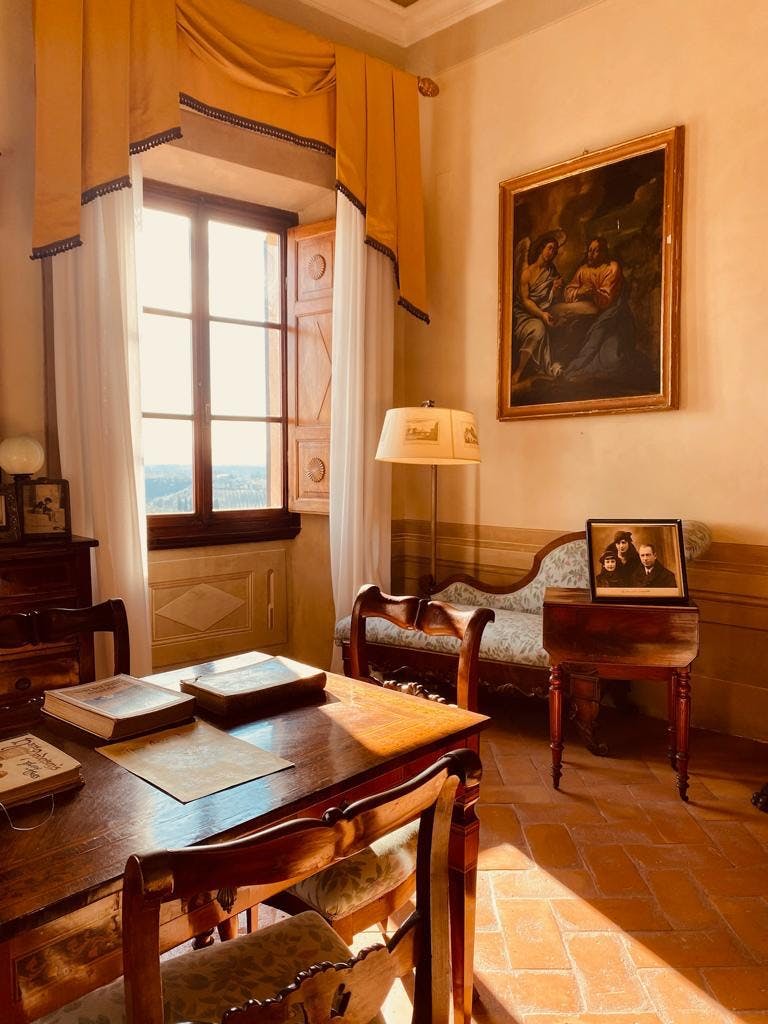 Un'accogliente sala studio con una scrivania, libri, poltrone, una lampada, una foto di famiglia incorniciata e un quadro, inondata dalla luce naturale di una finestra.
