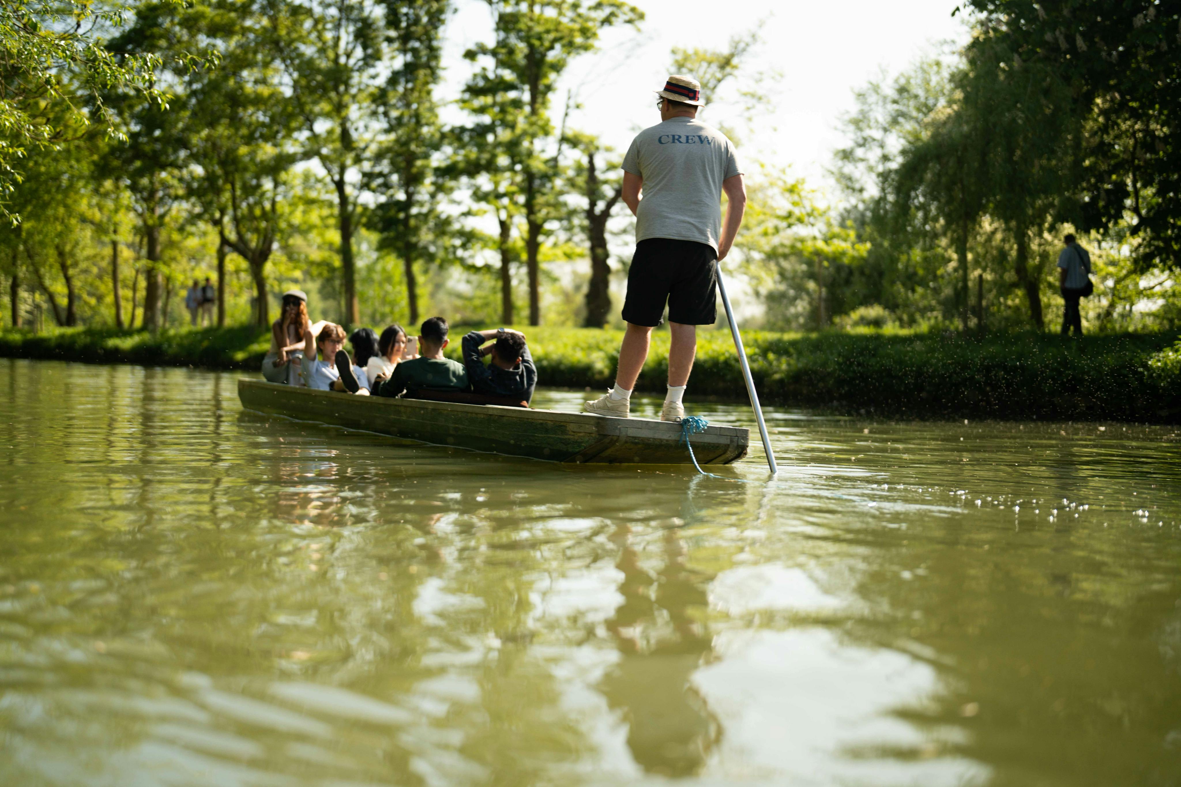 A person steers a small boat filled with seated passengers through a calm, tree-lined waterway on a sunny day.