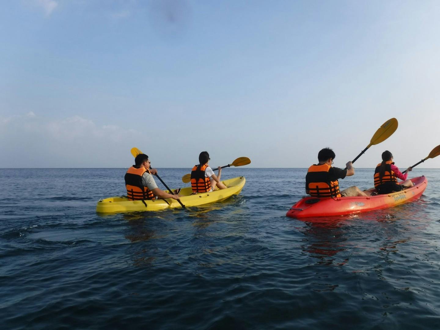 Three people wearing life jackets paddle kayaks on calm water under a clear sky.
