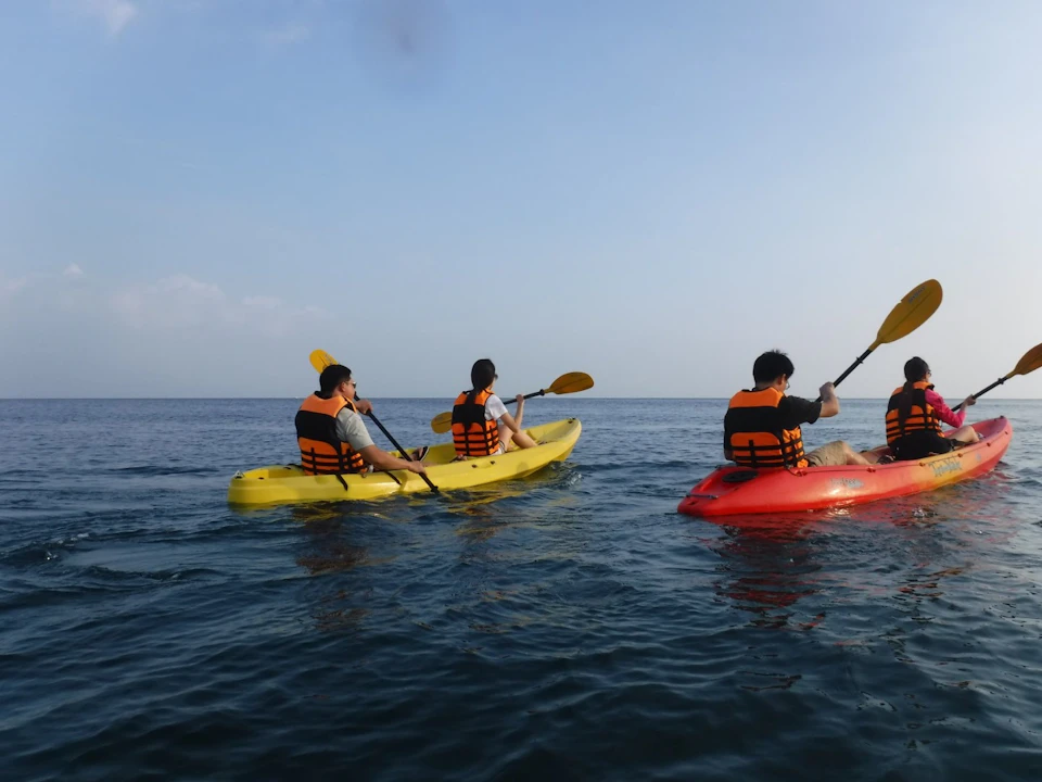 Three people wearing life jackets paddle kayaks on calm water under a clear sky.