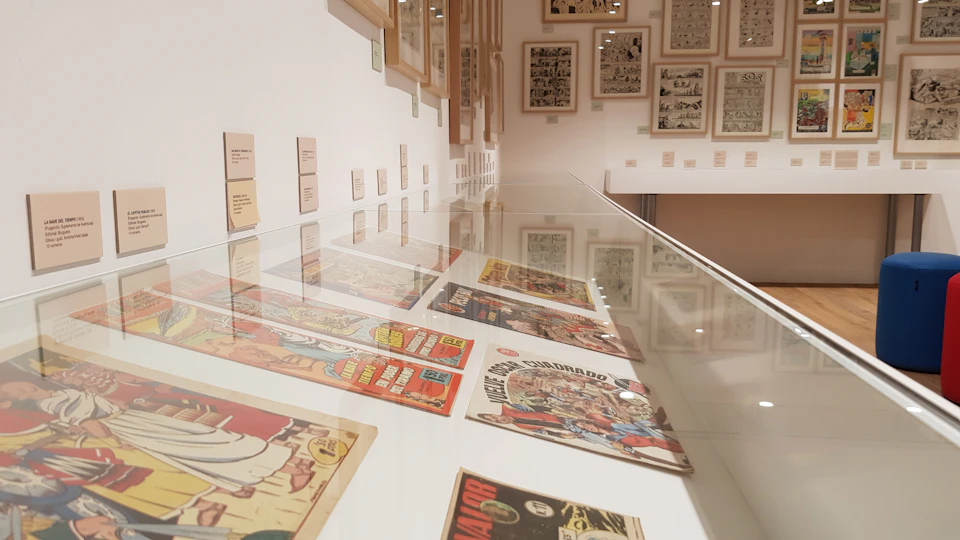 Comic books displayed in a glass case with framed comic pages and descriptions on the walls in a well-lit exhibition room.