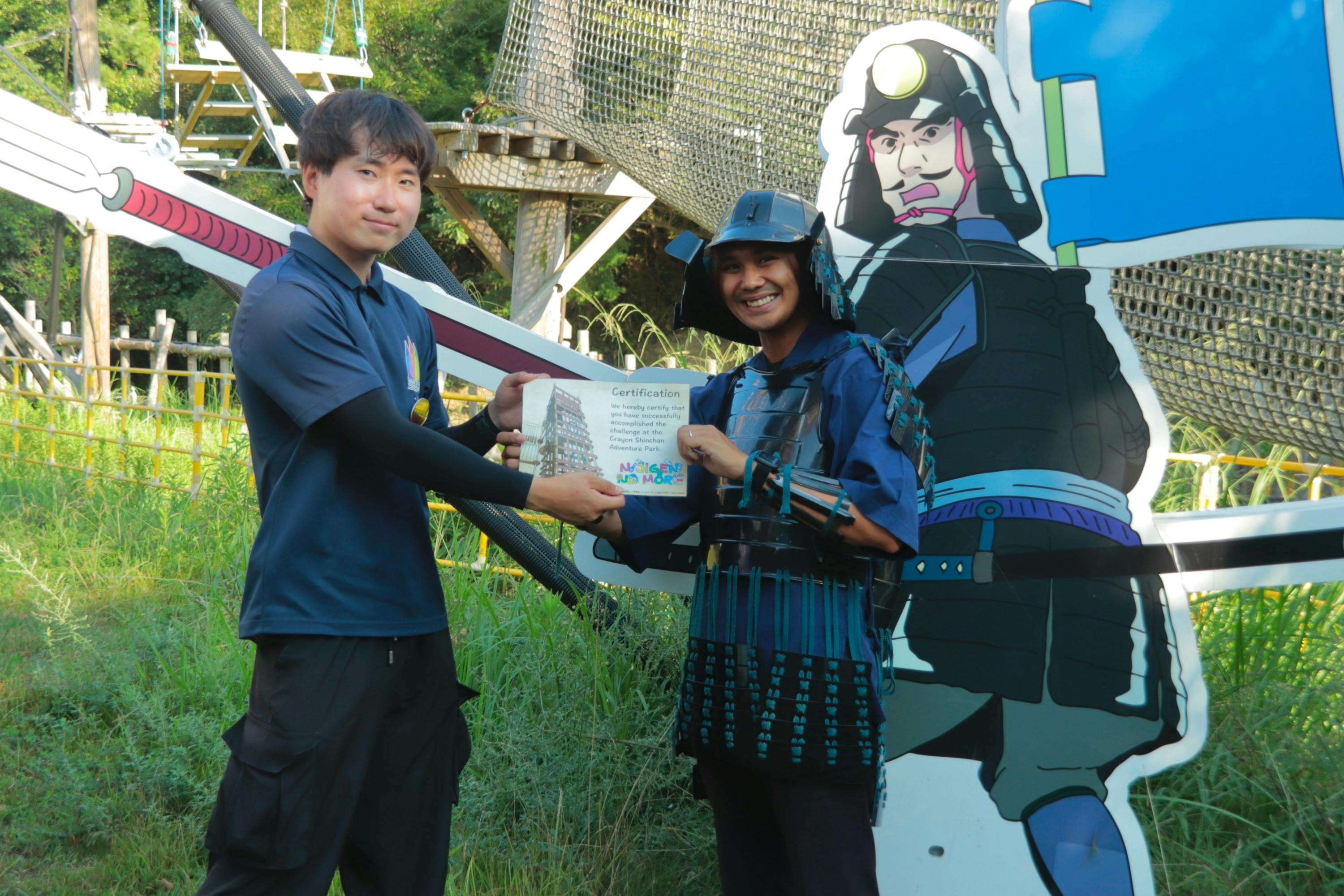 Two individuals standing outdoors; one presents a certificate to the other, who is dressed in a samurai-themed outfit.