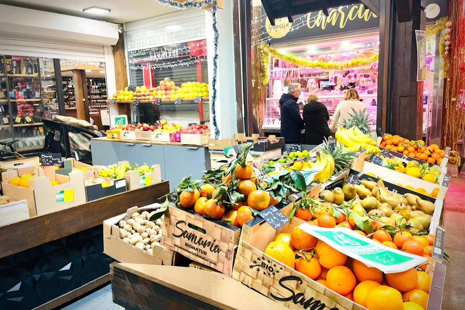 A market stall displaying various fruits like oranges, bananas, and ginger, with people shopping in the background.