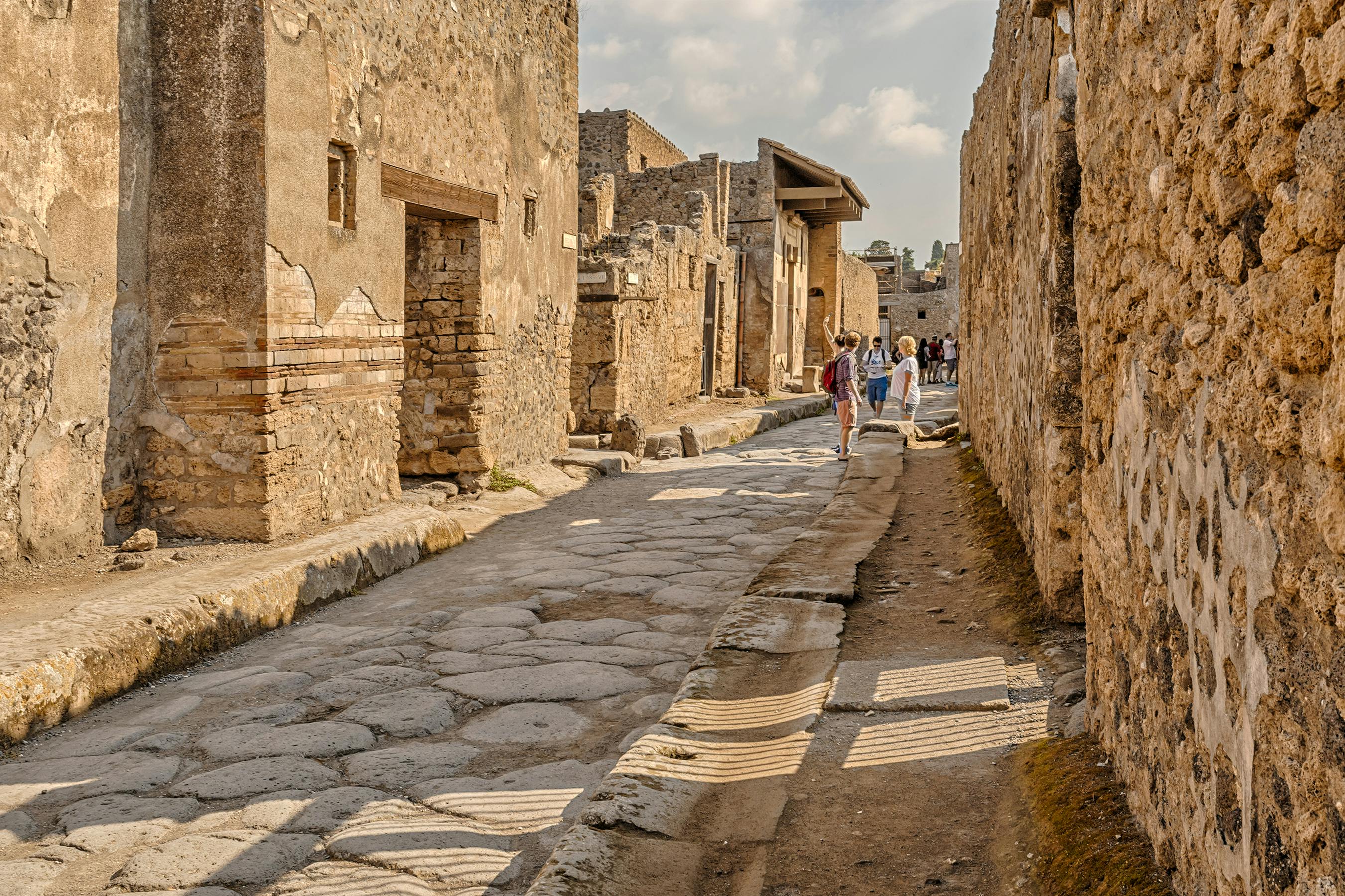 Ancient stone street lined with ruins, people walking, cobblestone path, sunny sky, shadows cast on walls and ground.