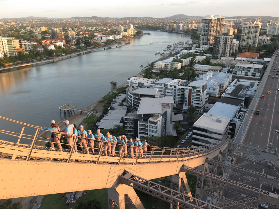 A group of people in blue shirts walk on a high bridge overlooking a river, with city buildings and hills in the background.