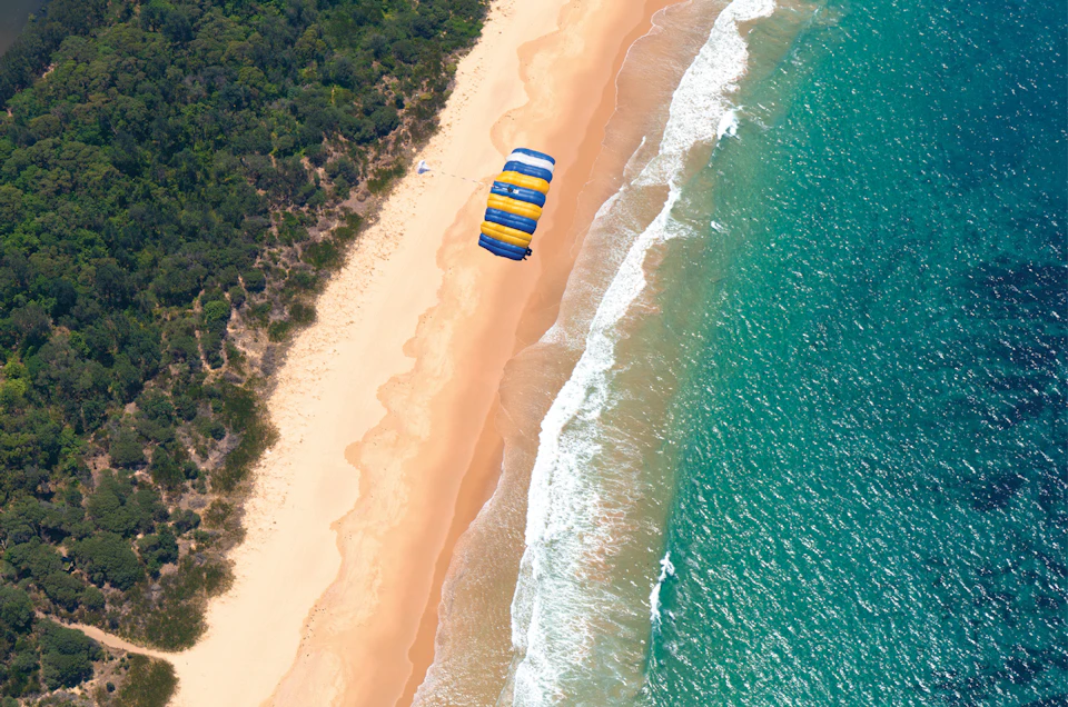 Aerial view of a person parasailing with a yellow and blue parachute over a sandy beach and turquoise ocean.